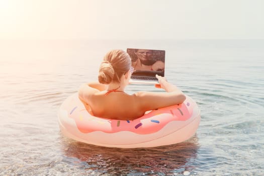 Woman freelancer works on laptop swimming in sea on pink inflatable ring. Pretty lady typing on computer while floating in the sea on inflatable donut at sunset. Freelance, remote work on vacation