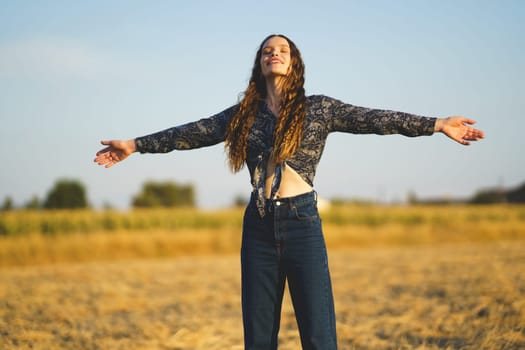 A young woman stands in a vast, golden field with her arms outstretched wide, fully embracing the warmth of the sun and the stunning beauty of nature surrounding her, feeling truly alive and free