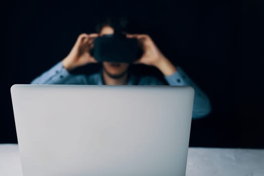 Blurred man in VR headset operating laptop in dark setting, depicting virtual reality technology use.