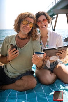 Middle-aged man and woman sitting on the beach with surprised face, browsing smartphone apps. looking at camera Concept:Holidays y Technology