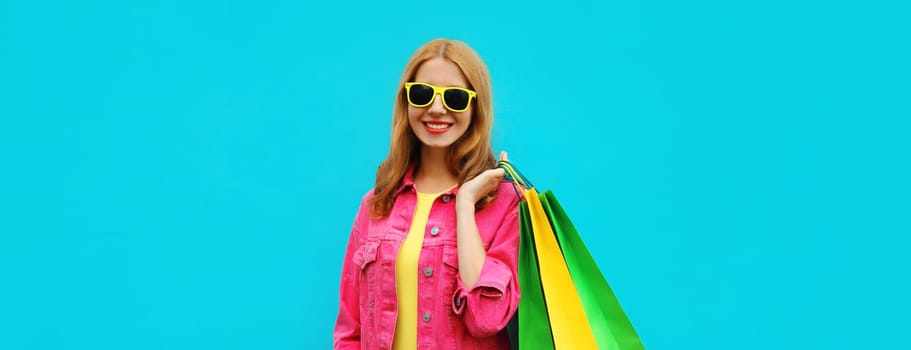 Shopping day, stylish beautiful happy smiling young woman posing with colorful shopping bags wearing bright pink clothing, summer glasses on blue studio background