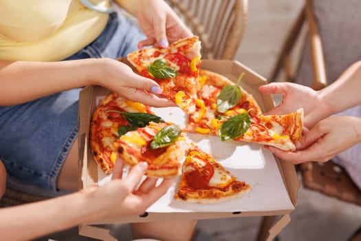 A group of friends share pizza outdoors, hands reaching for slices garnished with fresh basil, as a pizza box and plates of appetizers sit in the background.