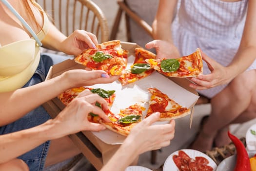 A group of friends share pizza outdoors, hands reaching for slices garnished with fresh basil, as a pizza box and plates of appetizers sit in the background.