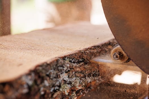A close-up view of a log being cut on a sawmill. The saw blade is visible, and sawdust is flying around, indicating active cutting. The log shows natural wood grain and a rough texture.