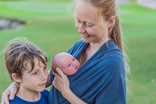 Mom walks with a newborn in a sling and also with her teenage son. This scene showcases the bond within the family, highlighting the nurturing care of a mother and the connection between siblings.