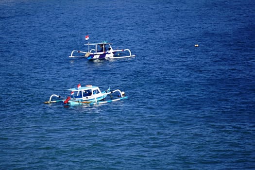 Two traditional Indonesian fishing boats with distinctive outriggers are anchored in the calm, blue waters of Pantai Senggigi, Lombok, Indonesia, with the Indonesian flag proudly flying on each.