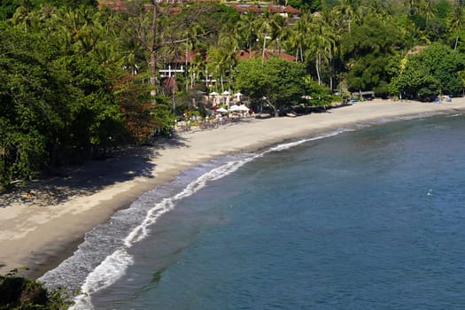Idyllic beach scene near Pantai Senggigi, Lombok, Indonesia, featuring pristine white sand, gentle waves, lush greenery, and a beachfront resort with palm trees.