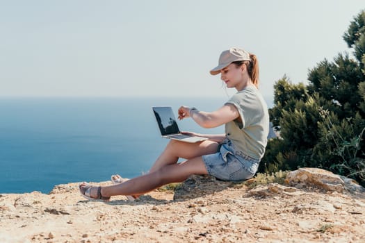 A young woman wearing a hat and casual clothes uses a laptop on a cliff with ocean views.