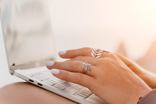 Digital nomad, woman in the hat, a business woman with a laptop sits on the rocks by the sea during sunset, makes a business transaction online from a distance. Freelance, remote work on vacation.