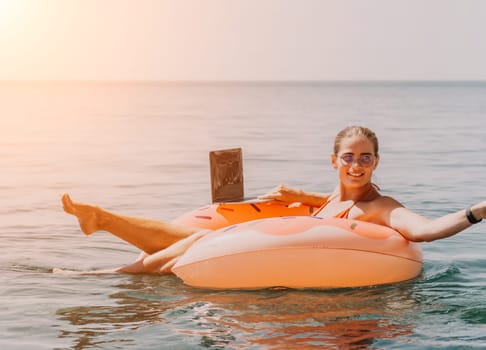 Woman freelancer works on laptop swimming in sea on pink inflatable ring. Pretty lady typing on computer while floating in the sea on inflatable donut at sunset. Freelance, remote work on vacation