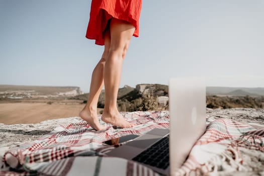 A woman in a red dress stands on a blanket with a laptop on it. The backdrop is a rocky cliff.