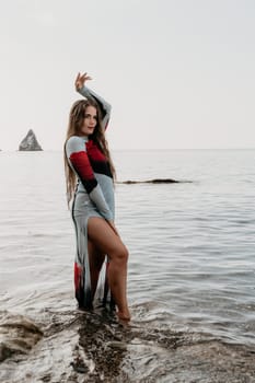 A woman in a colorful dress stands in shallow water, one hand raised, gazing towards a distant rock formation.