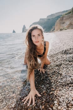 A woman with long brown hair poses on a beach covered in pebbles, with the ocean and a rocky cliff behind her.
