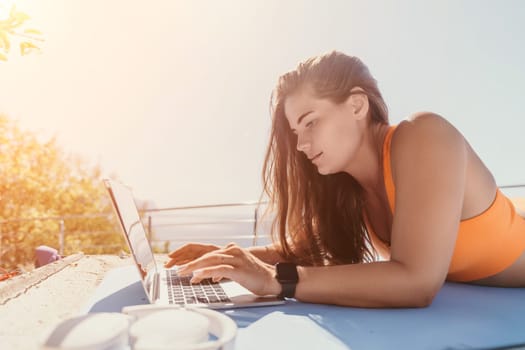 A woman with long brown hair is working on a laptop computer while laying on a blue mat outside.