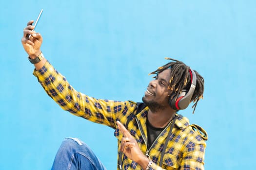 young black man with headphones taking a selfie with his smart phone with a blue wall in the background, technology and lifestyle concept, copy space for text