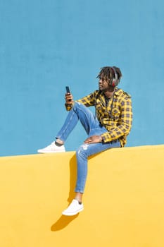 young black man sitting on a yellow wall with headphones consulting his smart phone with a blue wall in the background, technology and lifestyle concept, copy space for text