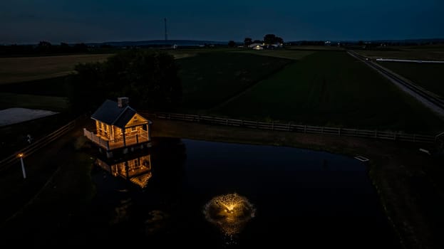 Tranquil Farm Scene At Dusk Featuring A Cozy Stone House Illuminated By Soft Lighting, Reflecting In A Peaceful Pond With A Fountain, Surrounded By Expansive Green Fields
