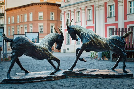 An artistic depiction of two bronze goat statues in a dynamic pose, located in the historic city square of Poznan, Poland. These sculptures symbolize the famous Poznan goats, a well known cultural emblem of the city. The statues are set against the backdrop of classic European architecture, adding to the charm of this heritage site. Ideal for themes related to travel, culture, history, and European landmarks