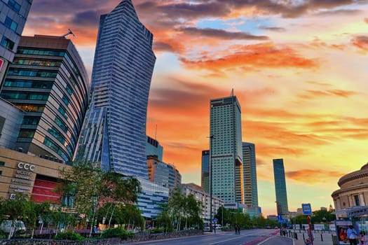 A stunning image of Warsaw's skyline featuring modern skyscrapers against a colorful sunset sky. The dynamic architecture, including curved and towering buildings, highlights the contemporary urban development of Poland capital. The warm hues of the sky contrast beautifully with the sleek designs of the buildings, creating an eye-catching cityscape. Ideal for use in travel, architecture, and cityscape themes.
