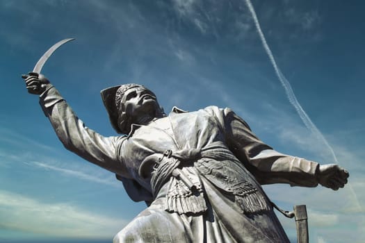 A striking image of a historic statue of a warrior brandishing a sword against a vivid blue sky. The powerful stance and detailed craftsmanship of the statue convey strength and determination, making it a prominent cultural landmark. Captured with a low-angle perspective, the statue symbolizes bravery and heroism. Perfect for use in historical, cultural, and travel-related themes.