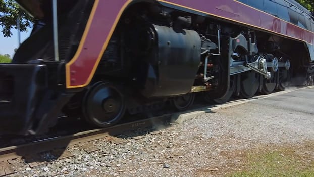 A vintage steam locomotive rolls by on the tracks, with heavy wheels turning and sunlight illuminating the scene. Gravel and grass line the railways, creating a rustic atmosphere.
