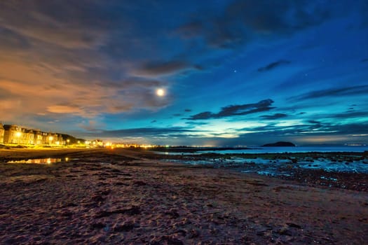 A breathtaking view of North Berwick Beach in the United Kingdom captured during dusk. The image showcases a vibrant sky transitioning into night with shades of blue, orange, and purple, while city lights illuminate the shoreline in the distance. The calm sea reflects the evening sky, creating a serene and captivating scene. Perfect for themes related to coastal landscapes, twilight photography, and travel.