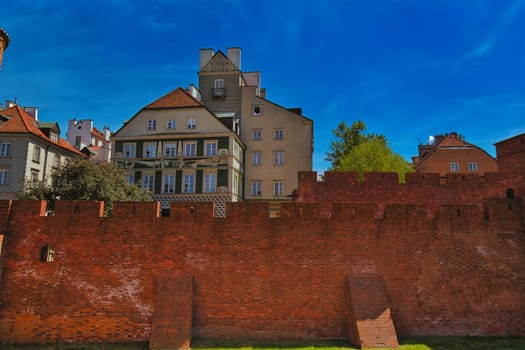 A stunning view of the historic brick walls in Warsaw Old Town, with traditional European buildings in the background. The well-preserved defensive walls are part of Warsaw rich history and architectural heritage, representing the city resilience through centuries. The vibrant blue sky contrasts with the red bricks, making this scene perfect for use in travel, history, and architecture themes.