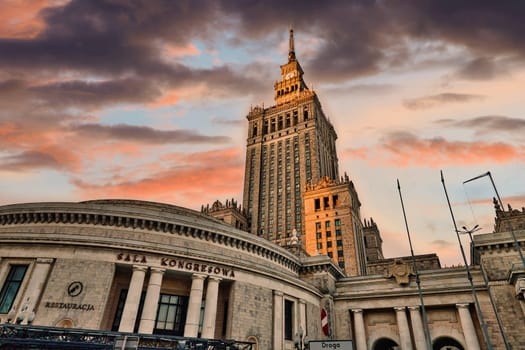 A striking image of the Palace of Culture and Science in Warsaw, Poland, captured at sunset. The towering structure, a symbol of post-war architecture, dominates the skyline with its impressive design and intricate details. The colorful sky adds drama and contrast, making the iconic building stand out even more. Perfect for themes related to travel, history, and urban architecture.
