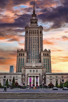 A majestic front view of the Palace of Culture and Science in Warsaw, Poland, taken during sunset. The building, an iconic symbol of Warsaw, stands tall with its grand architecture and historical significance, built in the 1950s as a gift from the Soviet Union. The vivid colors of the sky create a stunning contrast against the stone facade, making it a striking image of one of Warsaw most famous landmarks. Ideal for themes related to architecture, history, and travel.