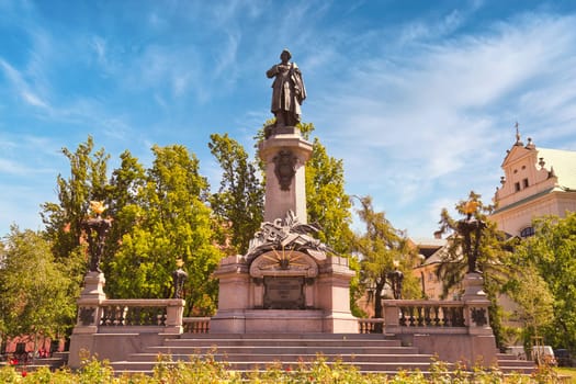 A grand view of the Adam Mickiewicz Monument located in Warsaw, Poland, set against a bright blue sky. This statue commemorates one of Poland greatest poets and is a significant cultural and historical landmark. Surrounded by lush greenery and intricate sculptures, the monument stands proudly in a public square, attracting visitors and locals alike. Ideal for themes related to history, culture, and travel.