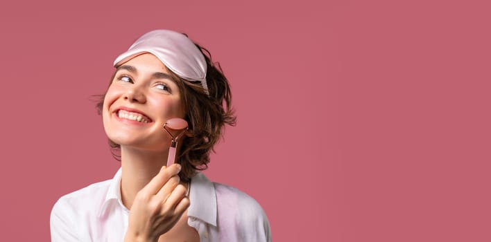 Young woman with rose quartz stone roller on pink background. Facial self care, beauty rituals, cosmetology, anti aging and anti-wrinkle treatment. Copy space. High quality