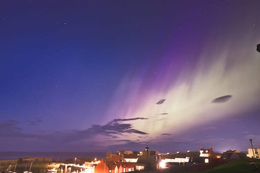 A captivating image of the Aurora Borealis illuminating the sky over a coastal town in North Berwick, Scotland. The ethereal green and purple auroras stretch across the night sky, casting a magical glow over the distant rooftops and calm sea. This rare natural phenomenon beautifully contrasts with the town twinkling lights, creating a breathtaking scene of nature meeting civilization. Ideal for themes related to nature, night sky photography, and travel
