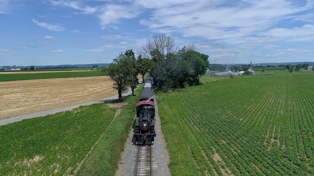 A vintage steam train chugs along the tracks, surrounded by lush green fields and trees, creating a scenic view of rural life on a bright day.
