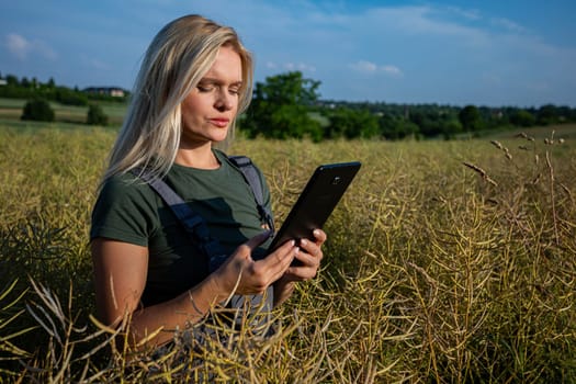 A woman farmer is checking the quality of rapeseed on the middle of the field and holding a tablet. She is checking the information about her harvest and she seems worried about the quality of rapeseed. She owns a rapeseed field and is checking this year's harvest. She is an experienced farmer, caring for her crops in a vast field. She has blonde hair. The soil is fertile and productive. The rapeseed is yellow and ready for harvest. Satisfied with the bountiful and lavish harvest, the woman farmer, adorned in working clothes and with tanned skin, gazes upon the expansive yellow landscape with a sense of fulfillment. With the blue sky adorned with fluffy white clouds overhead, she stands in the middle of the field, her spirit merging with the warmth of the sun, celebrating the culmination of her dedication and the richness of the land's yield.