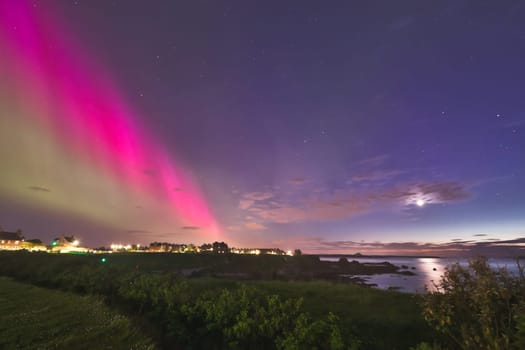 A stunning capture of the Aurora Borealis lighting up the sky over North Berwick coastline. The striking pink and green auroras contrast beautifully with the darkening sky and calm coastal waters, creating a serene yet dramatic scene. The town lights in the distance, along with the crescent moon, add to the ethereal beauty of this rare phenomenon. Perfect for use in travel, nature, and astrophotography themes.