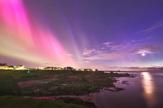 An awe-inspiring capture of the Aurora Borealis illuminating the sky over North Berwick, Scotland. The vibrant pinks and purples of the aurora stretch across the horizon, reflecting beautifully on the calm coastal waters. The scene is further enhanced by the moonlight and the distant lights of the coastal town, creating a serene yet magical atmosphere. Perfect for themes related to nature, travel, and night sky photography.