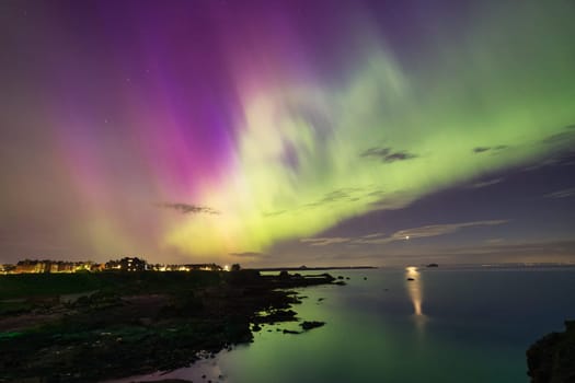 An enchanting image of the Aurora Borealis lighting up the night sky over the calm waters of North Berwick, Scotland. The vivid green and purple lights stretch across the sky, reflecting beautifully on the sea below. The tranquil coastal landscape and distant lights enhance the magical atmosphere of this rare natural phenomenon. Perfect for themes related to nature, night photography, and travel.