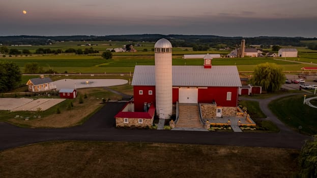 Ronks, Pennsylvania, USA, July 19 2024 - Peaceful Farm Scene At Dusk Featuring A Large Red Barn With A Prominent White Silo, A Stone Pathway, And Surrounding Buildings, All Set Against A Backdrop Of Rolling Green Fields And A Softly Lit Sky.