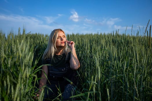 In the tranquil expanse of a rustic wheat field, a female farmer squats, holding a wheat stalk in one hand, contemplatively placing it near her lips. With sun-kissed skin and clad in work attire, she embodies the essence of rural life. Her expression is one of introspection, leaving viewers to ponder whether she is content with the harvest or harbors concerns. She is looking at the horizon. The vibrant green hue of the wheat stalk contrasts against the golden tones of the field, adding depth to the scene. Against the backdrop of the sprawling countryside, her presence evokes a sense of agricultural tradition and connection to the land. This image captures the quiet moments of farming, where contemplation mingles with the labor of the harvest. As she gazes thoughtfully upon the wheat, her blonde hair softly frames her face, enhancing the rustic charm of the scene