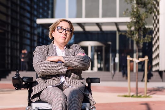 businesswoman using wheelchair with arms crossed in front of the entrance of an office building in the financial district, concept of diversity and urban lifestyle, copy space for text