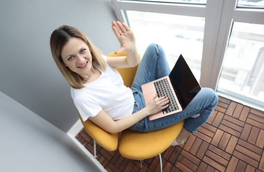 Young woman with laptop sitting in armchair and waving hand. Telework concept