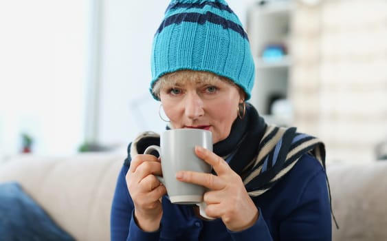 Portrait of an elderly woman in warm hat holding cup with medicine. Colds in elderly concept