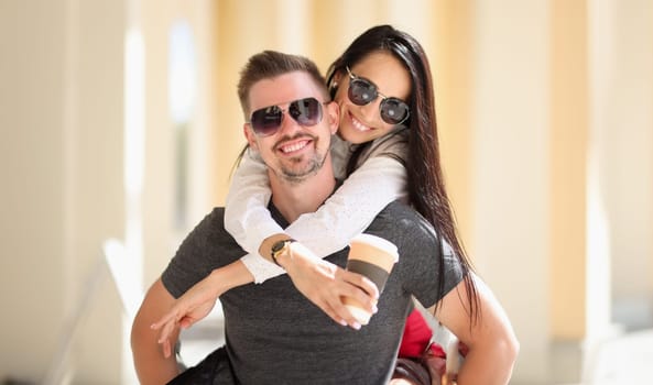 Smiling woman in sunglasses holds coffee and hugs man. Happy harmonious relationship concept