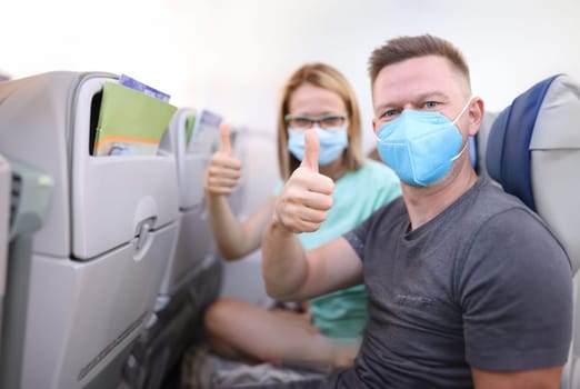 Man and woman in medical protective masks in aircraft cabin hold their thumbs up. Safe flights to coronavirus pandemic concept