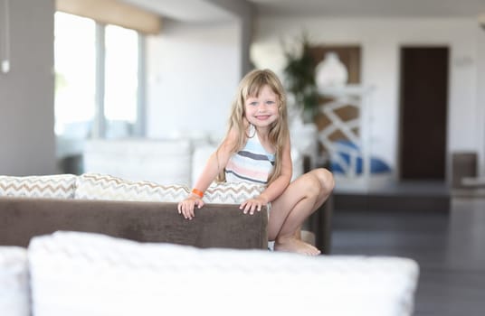 Little smiling girl sits with her legs on couch. Happy and carefree childhood concept