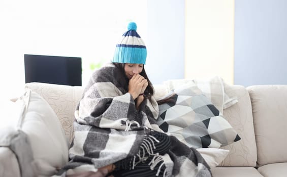 Frozen woman in hat and blanket sits on couch. Heating in houses and apartments during the cold season concept