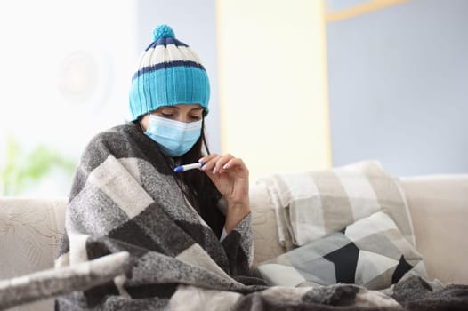Frozen woman in hat and blankets in protective mask sits on sofa with thermometer. Seasonal diseases and coronavirus pandemic concept