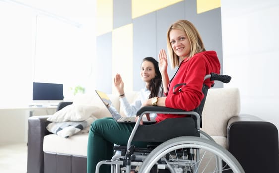 Woman in wheelchair with laptop on her lap waving in greeting with her girlfriend. Remote work for people with disabilities health concept