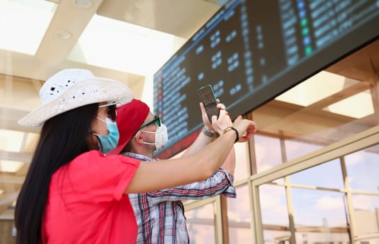 Woman and man wearing medical protective masks take pictures of an information board at train station. Travel to coronavirus pandemic concept