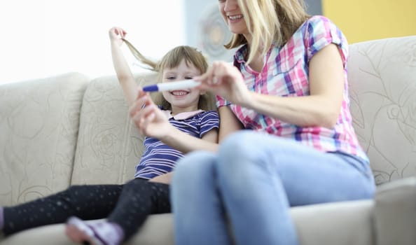 Joyful mom and daughter are sitting on couch and holding pregnancy test. How to prepare child for new addition to the family concept